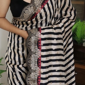 May include: A black and white striped saree with a decorative border. The saree features a black sleeveless blouse. The border has a floral design with gold accents and a red trim. The saree has the text "DE" and "RPL-RM-0063".