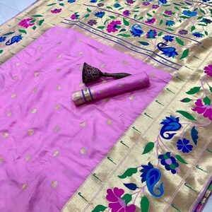 May include: A pink sari with gold floral borders and accents. The sari features a rolled-up section and a decorative object. The gold border has a floral design with blue, pink, and green accents. The sari has a gold and pink color scheme.