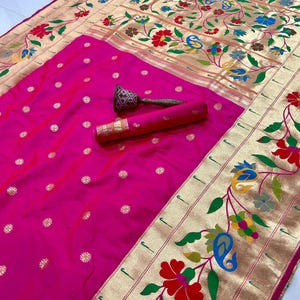 May include: A vibrant pink saree with gold floral and paisley border details. The saree features gold accents and small gold floral patterns. A rolled-up section of the saree and a decorative item are visible.