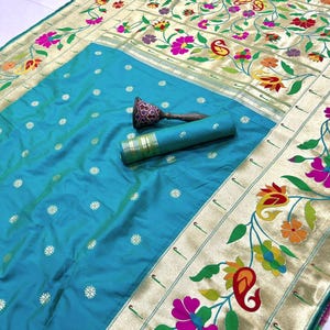 May include: A vibrant turquoise saree with a gold border featuring floral and paisley designs in pink, yellow, and green. The saree is folded and rolled, with a decorative handle on top. The fabric has small white floral accents.