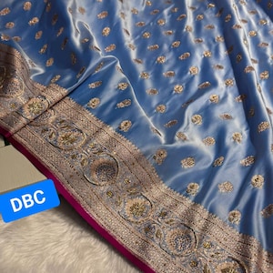 May include: A pale blue fabric with gold detailing and an ornate border. The fabric has a soft, flowing texture and is decorated with small, scattered gold accents. The border features intricate embroidery and a bright pink trim. The text "DBC" is visible in a blue rectangle.