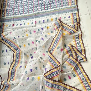 May include: White textile with a repeating floral pattern in pink, purple, yellow, and green. The border features a detailed design with similar colors and a brown trim. The fabric is draped, showcasing its texture and design.