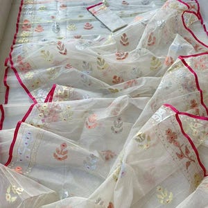 May include: A white, sheer fabric with intricate floral and geometric patterns in shades of pink, gold, and green. The edges are trimmed with a vibrant pink border. A matching fabric piece is also included.