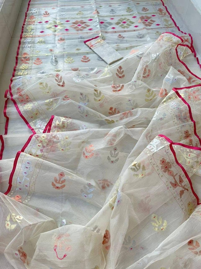 May include: A white, sheer fabric with intricate floral and geometric patterns in shades of pink, gold, and green. The edges are trimmed with a vibrant pink border. A matching fabric piece is also included.