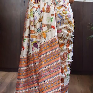 May include: A white saree with a colorful bird pattern and an orange and pink geometric border. The saree has a decorative fringe and the text "DBC" and "KTH-LMB-150925" in a white box.