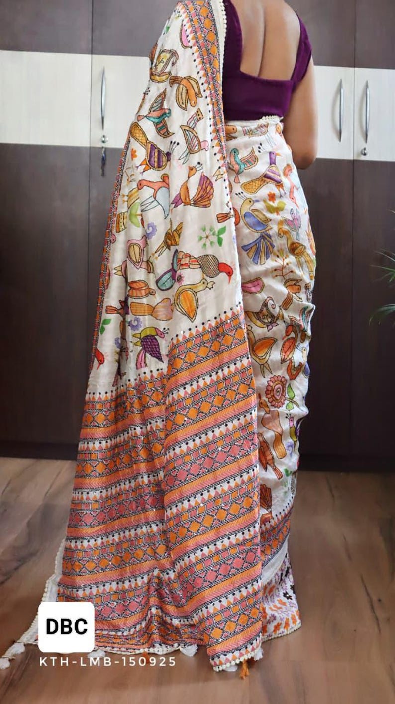 May include: A white saree with a colorful bird pattern and an orange and pink geometric border. The saree has a decorative fringe and the text "DBC" and "KTH-LMB-150925" in a white box.