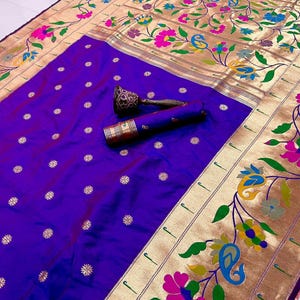 May include: A vibrant purple saree with a gold border featuring floral and paisley designs in pink, blue, and green. The saree has small, gold floral accents and a rolled-up section with decorative ends.