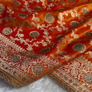 May include: Close-up of a vibrant orange and red fabric with intricate gold embroidery. The design features floral patterns and detailed borders, creating a luxurious and ornate appearance. The fabric has a silky sheen.