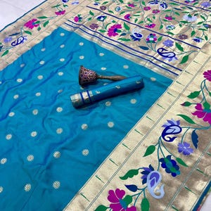 May include: A vibrant turquoise silk saree with intricate floral embroidery in pink, purple, and blue. The saree features a gold border with detailed patterns and a matching rolled fabric piece. A decorative handle rests on the fabric.