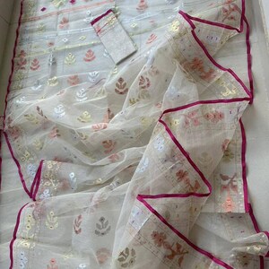 May include: A sheer white saree with intricate floral and geometric patterns in gold, pink, and silver. The saree has a vibrant pink border and a matching blouse piece. This traditional garment is perfect for special occasions.