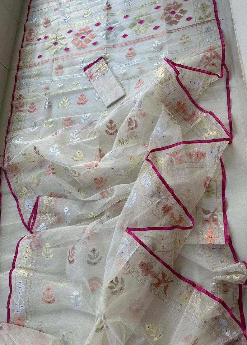 May include: A sheer white saree with intricate floral and geometric patterns in gold, pink, and silver. The saree has a vibrant pink border and a matching blouse piece. This traditional garment is perfect for special occasions.