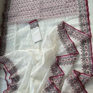 May include: White saree with a floral pattern and a matching blouse piece. The saree features a detailed border with burgundy and green accents. The fabric appears lightweight and semi-transparent, with a delicate texture. The overall design is elegant and traditional.