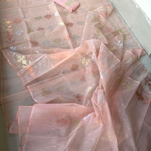 May include: A light peach-colored saree with intricate floral and geometric patterns. The sheer fabric drapes elegantly, with a matching rectangular piece folded on top. The saree has a delicate, shimmering appearance.