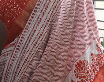 May include: A saree featuring a white base with red chevron patterns and floral borders. The fabric includes a solid red section with white geometric prints and a metallic gold border. A silver necklace is visible at the neckline. Text reads DBC KTH-MNG-230726.