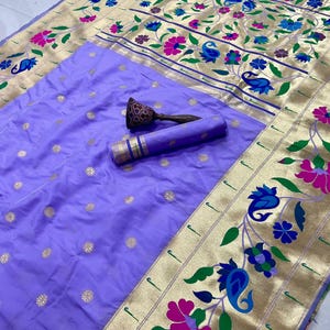 May include: A lavender-colored sari with gold floral embroidery and a decorative border featuring pink, blue, and green floral patterns. A rolled-up section of the sari and a decorative wooden object are also visible.