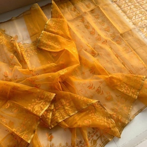May include: A sheer, golden-yellow fabric with intricate floral and paisley patterns. The fabric is draped, revealing layers and textures. The edges are trimmed with a gold border. The overall effect is delicate and luxurious.
