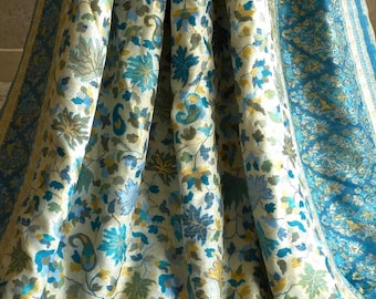 Può includere: Un tessuto drappeggiato con una base color crema, ornato da un motivo floreale dettagliato nei toni del blu, verde e giallo. Il design include fiori stilizzati e motivi paisley, creando un aspetto ricco e strutturato. È visibile un bordo blu con un motivo simile.