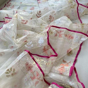 May include: A sheer, off-white fabric with floral patterns in pink, gold, and green. The edges are trimmed with a vibrant fuchsia border. The delicate material is draped, showcasing its lightweight quality and intricate design.