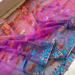 May include: A sheer, purple saree with intricate floral embroidery in orange, blue, and pink. The fabric transitions from a lighter pink to a deeper purple, with detailed borders. The saree is folded, showcasing its delicate texture and vibrant colors.