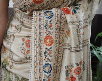 May include: Cream-colored saree with intricate floral patterns in orange, blue, and green. The saree features detailed embroidery and a delicate lace trim along the border. The text "KTW-PSLY-140125" is visible.