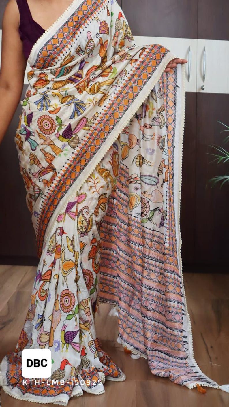 May include: White saree with a colorful bird and floral pattern. The saree has an orange and white border with a decorative trim. The text "DBC" and "KTH-LMB-15092" are visible on the saree.