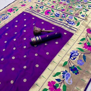 May include: A vibrant purple sari with gold borders and floral embroidery. The sari features a detailed design with pink, blue, and green flowers, and the word "Jambali" is visible in the top left corner.