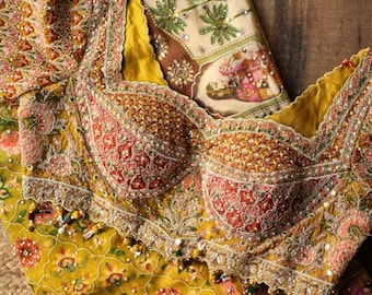 May include: An ornate, embroidered bodice in shades of gold, red, and pink, with intricate beadwork and a scalloped neckline. The garment is set against a backdrop of patterned fabric in complementary colors, suggesting a traditional or cultural design.