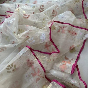 May include: A sheer, off-white fabric with a floral pattern in shades of pink, gold, and green. The edges are trimmed with a vibrant fuchsia border. The fabric is draped, showcasing its delicate texture and design.