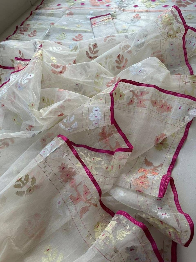 May include: A sheer, off-white fabric with a floral pattern in shades of pink, gold, and green. The edges are trimmed with a vibrant fuchsia border. The fabric is draped, showcasing its delicate texture and design.