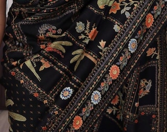 May include: Black shawl with intricate floral and botanical embroidery in orange, blue, and green. The design features detailed patterns and a border with a repeating floral motif. The shawl is draped, showcasing the detailed craftsmanship.