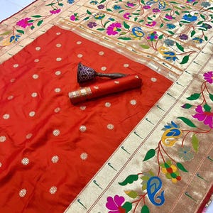 May include: An orange sari with a floral and paisley design. The border features gold trim and colorful flowers. The word "Orange" is in the top left corner. A decorative item is on the sari.