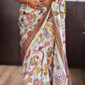 May include: White sari with a colorful, intricate pattern of birds, flowers, and other designs. The sari has orange and pink borders with a cream-colored trim. The text "DBC" is visible in the lower left corner.