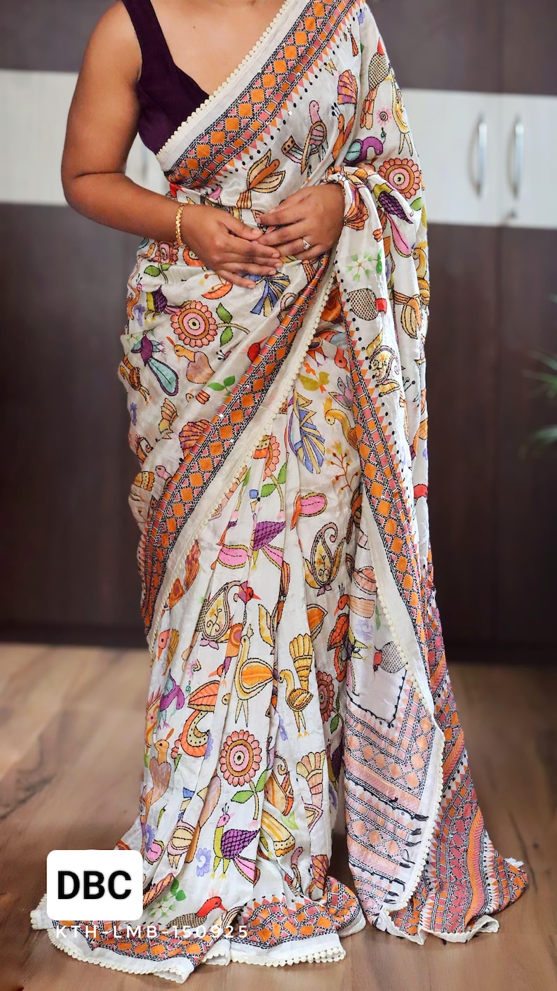 May include: White sari with a colorful, intricate pattern of birds, flowers, and other designs. The sari has orange and pink borders with a cream-colored trim. The text "DBC" is visible in the lower left corner.