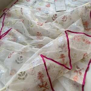May include: A white sheer fabric with floral and leaf patterns in pastel colors. The fabric has a decorative border with pink trim. The fabric is draped and folded, suggesting it is a garment or textile.