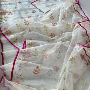May include: A white fabric ensemble with intricate floral and geometric patterns in shades of pink, gold, and silver. The fabric has a sheer, flowing quality, with a vibrant pink border. The set includes a matching top and scarf.