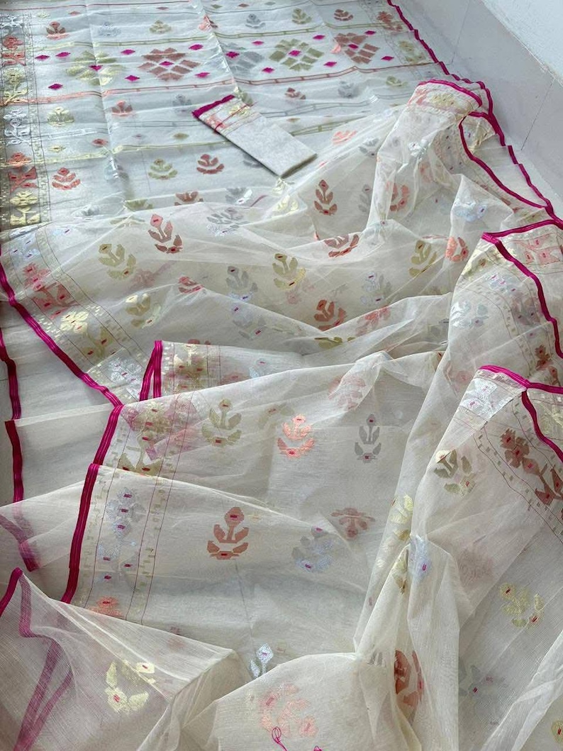 May include: A white fabric ensemble with intricate floral and geometric patterns in shades of pink, gold, and silver. The fabric has a sheer, flowing quality, with a vibrant pink border. The set includes a matching top and scarf.