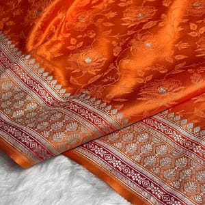 Puede incluir: Primer plano de una tela naranja vibrante con un estampado floral. La tela tiene un borde detallado con detalles plateados y rojos. El material está doblado, mostrando su textura y diseño.