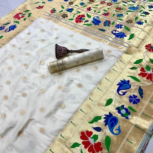 May include: A white sari with gold floral accents and a decorative border featuring red, blue, and green floral patterns. A rolled-up fabric and a decorative object are also present.