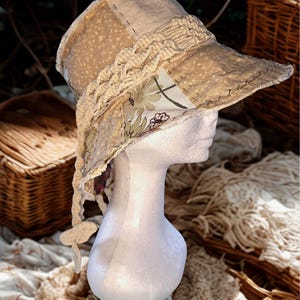 May include: A beige patchwork sun hat with a wide brim, featuring floral and dotted fabric panels. The hat has a braided trim and a chin strap. The text "Celebrate what sets you apart" is visible in the upper left corner. The hat is displayed on a mannequin head.