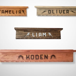 May include: Wooden shelves with names and sports-themed silhouettes. The shelves are in various wood tones, with names like "AMELIA", "OLIVER", "LIAM", and "KODEN" displayed. Each shelf has small pegs for hanging items.
