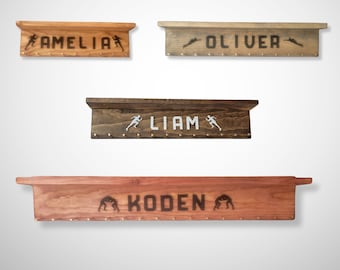 May include: Wooden shelves with names and sports-themed silhouettes. The shelves are in various wood tones, with names like "AMELIA", "OLIVER", "LIAM", and "KODEN" displayed. Each shelf has small pegs for hanging items.