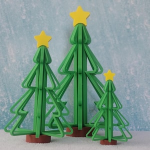 May include: Three green Christmas tree decorations of varying sizes, each topped with a yellow star. The trees have a geometric design and are set on brown bases. The background is a light blue with a snowy texture.