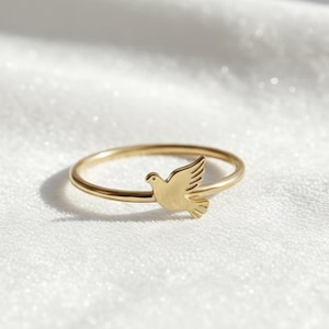 Op de afbeelding: Een delicate gouden ring met een duifontwerp. De ring heeft een dunne band en een klein, gedetailleerd duifsilhouet aan de voorkant. De ring wordt getoond op een wit, getextureerd oppervlak.
