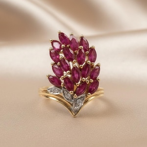 May include: A gold ring featuring a cluster of oval-cut, deep red gemstones, possibly rubies, arranged in a floral or leaf-like pattern. Small, clear accent stones are set near the base of the design. The ring sits on a satin surface.