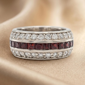 May include: A silver ring with three rows of gemstones. The top and bottom rows feature small, round, clear stones. The middle row is composed of square, deep red stones. The ring is set against a soft, golden background.
