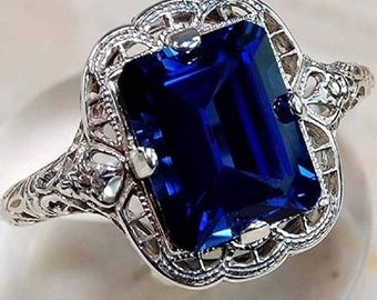 May include: A silver ring with a large, rectangular, deep blue gemstone. The ring has an ornate, filigree design around the stone and on the band. The gemstone is the focal point, catching the light and creating a striking visual.