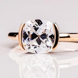 May include: A close-up of a gold ring featuring a large, square-cut diamond. The diamond is clear and multifaceted, reflecting light. The ring has a simple, elegant design, with the diamond set in a gold bezel.