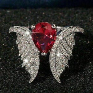 May include: A silver-toned ring featuring a large, pear-shaped, red gemstone at the center. The ring's design includes two wings encrusted with small, sparkling clear stones, creating a glamorous and elegant look. The ring is set against a dark background.
