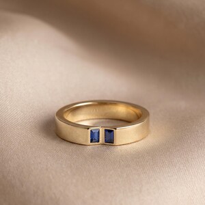May include: A gold band ring featuring two small, rectangular, blue sapphire gemstones set side-by-side in a channel setting. The ring has a smooth, polished finish and a flat profile, showcasing a clean and simple design with two vibrant blue stones.