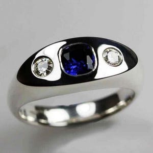 May include: A silver ring with a polished finish, featuring a large, square, deep blue gemstone in the center, flanked by two smaller, round, clear gemstones. The ring has a smooth, rounded band.